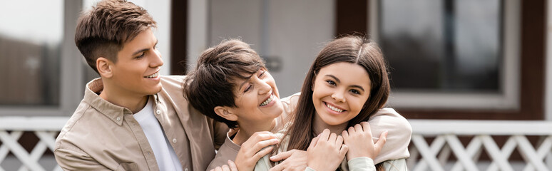 happy parents day, mother hugging cheerful teenage daughter near young adult son on backyard of family house, celebration, bonding, moments to remember, modern parenting, banner