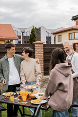 family grill party, celebration of parents day, happy young adult son hugging middle aged mother, bearded father in apron looking at teenage daughter, translation of tattoo: harm none do what you will