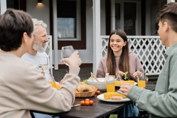 happy teenage girl looking at cheerful parents during family celebration, sitting on backyard of summer house, spending time together, eating grilled bbq food, happy parents day concept