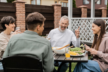 cheerful middle aged man looking at teenage daughter and gesturing during bbq party, sitting on backyard of summer house, spending time together, eating grilled bbq food, happy parents day concept
