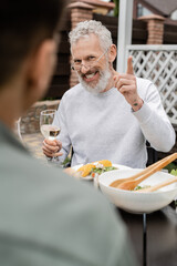 cheerful middle aged man looking son and gesturing during bbq party, blurred foreground, sitting on backyard of summer house, spending time together, eating grilled bbq food, parents day concept