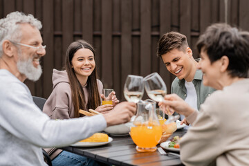 Smiling siblings sitting near tasty bbq food and blurred middle aged parents toasting with wine during parents day celebration at backyard, family love and unity concept, tradition and celebration