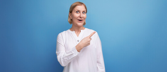 confident blond middle aged woman in white blouse pointing her finger to the side on studio background with copyspace