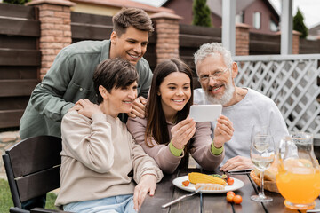 Smiling teen girl using smartphone near middle aged parents and brother during barbeque party with summer food and parents day celebration at backyard in june, happy parents day concept