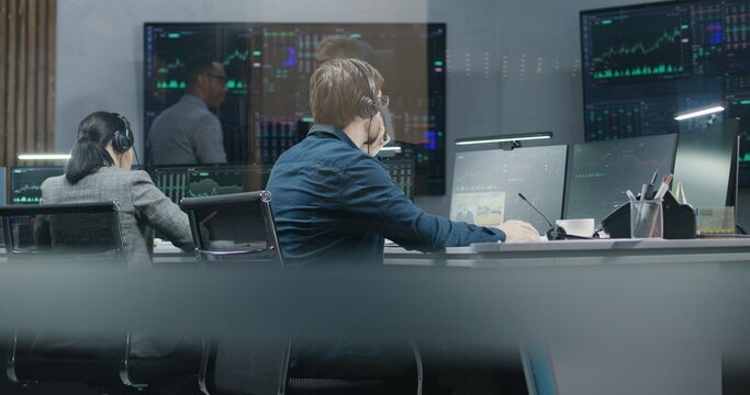 Diverse stock traders monitor real-time stocks data on computers, work in broker agency office. Colleagues watch exchange market charts on big digital screens. Cryptocurrency trading and investment.