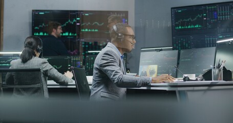 African American financial analyst monitors real-time stocks on computers, writes data with pen, works in bank office. Exchange market charts displayed on big digital screens. Investment or analytics.