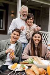 Cheerful mature parents hugging and looking at camera near children and summer food while celebrating parents day at backyard in june, special day for parents concept, tradition and celebration