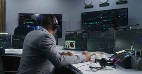 African American trader with pen in hand works at computer with displayed real-time stocks. Colleagues analyze exchange market charts on big screens at background. Concept of trading and investment.