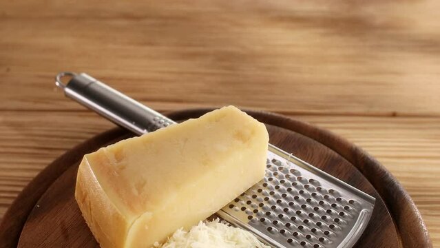 A piece of hard cheese and grated cheese with a grater on a cutting board. Wooden background. Traditional Italian cheese grana parmigiano reggiano. High quality 4k footage