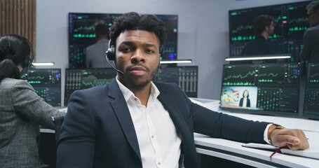 African American financial analyst works at computer with displayed real-time stocks, turns and looks at camera. Colleagues analyze exchange market charts at background. Investment concept. Portrait.
