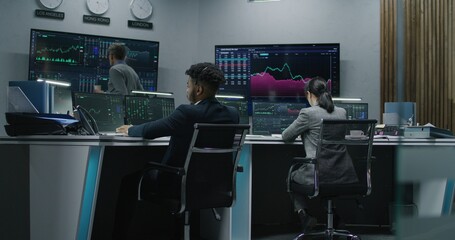 Multi ethnic team of traders work on multi-monitor computers with displayed real-time stocks. Colleagues analyze exchange market charts on multiple big screens. Cryptocurrency trading and investment.