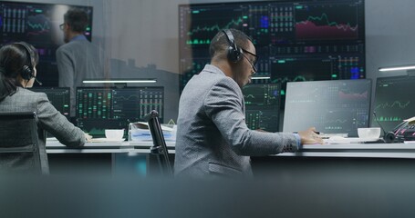 African American financial analyst monitors real-time stocks on computers, writes data with pen, works in bank office. Exchange market charts displayed on big digital screens. Investment or analytics.