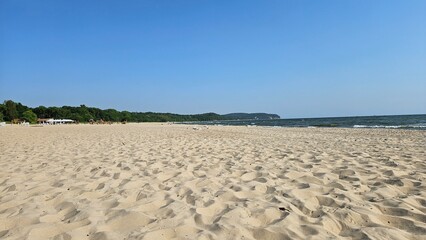 Ujmujące zdjęcie plaży, gdzie błękitne niebo harmonizuje z jasnym, delikatnym piaskiem. Promienie słońca rozświetlają krajobraz, tworząc atmosferę spokoju i sielanki. - obrazy, fototapety, plakaty