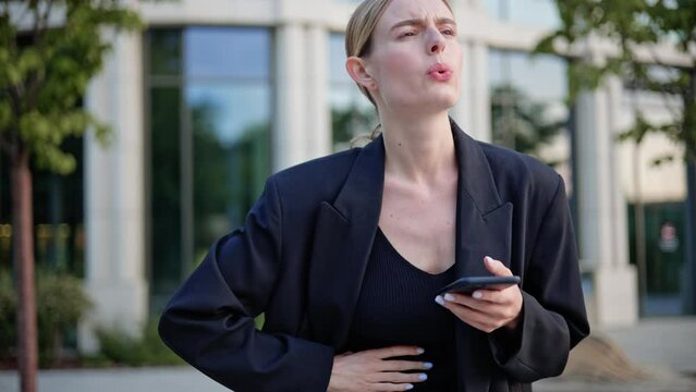 Unhappy woman feeling stomach pain, ache due to nervous before important event. Sick girl standing outdoor, holding belly, feeling abdominal or menstrual pain near office place holding mobile phone.