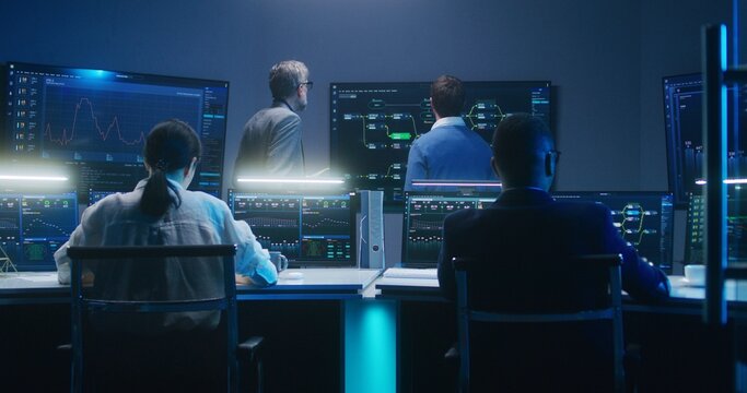 Multiethnic team of IT technical specialists and software programmers work with data server, analysis charts and blockchain network on computers and big digital screens in monitoring office at night.
