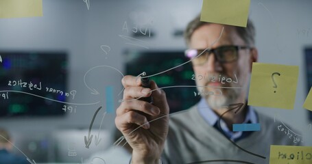 Mature financial analyst draws on glass wall with sticky notes in investment bank office. Businessman develops and analyzes business strategy for company. Colleagues work on computers on background.