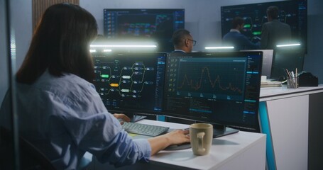 Female software engineer sits at computer with displayed blockchain network and surveillance maps. Technical support specialists work in monitoring room. Multiple big screens on the wall. Dolly shot.