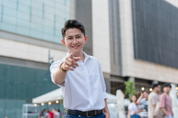 A handsome and confident asian man in his early 20s points to the camera. Outdoor city plaza scene.