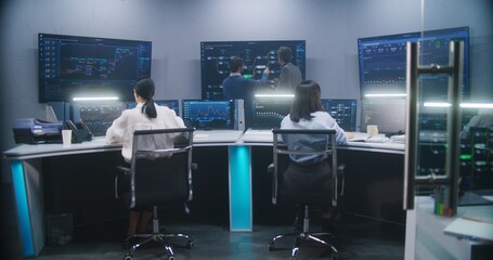 Female software engineer sits at computer with displayed blockchain network and surveillance maps. Technical support specialists work in monitoring room. Multiple big screens on the wall. Dolly shot.
