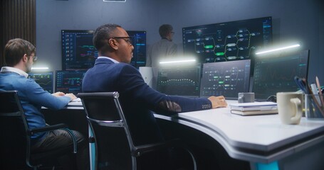 Multiethnic IT technical specialists sit in front of computers with data server in monitoring office. Software engineer works with live analysis charts and blockchain network on big digital screens.