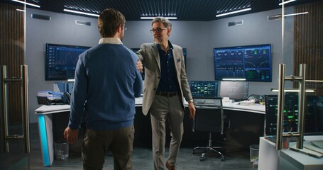 Mature supervisor in business suit greets programmers in monitoring room and looks at camera. Big data engineers start work. Multiple big screens with displayed real-time analysis charts. Dolly shot.