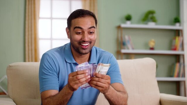 Happy excited indian man counting money or currency notes while sitting on sofa at home - concept of profit, won lottery and Financial success.
