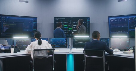 Female software engineer sits at computer with displayed blockchain network and surveillance maps. Technical support specialists work in monitoring room. Multiple big screens on the wall. Dolly shot.