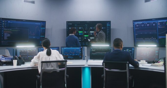 Female software engineer sits at computer with displayed blockchain network and surveillance maps. Technical support specialists work in monitoring room. Multiple big screens on the wall. Dolly shot.