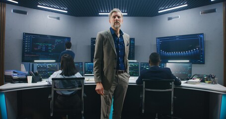 Mature big data scientist puts on glasses, stands in monitoring room and looks at camera. Software engineers work at computers at background. Multiple big screens with displayed live analysis feed.