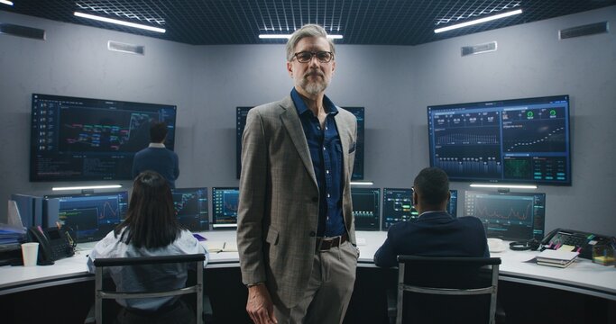 Mature big data scientist puts on glasses, stands in monitoring room and looks at camera. Software engineers work at computers at background. Multiple big screens with displayed live analysis feed.