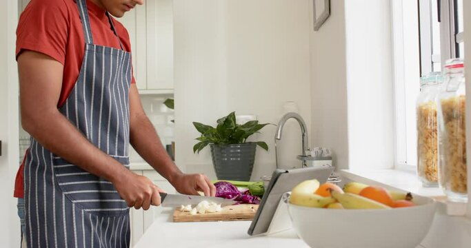 Biracial man in apron chopping vegetables and using tablet in kitchen, slow motion