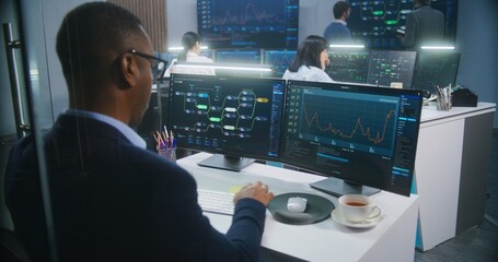 African American big data scientist works at computer in monitoring room. Multiracial colleagues analyze real-time charts at background. Multiple big screens on the wall with displayed server data.