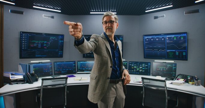 Mature supervisor in business suit greets programmers in monitoring room and looks at camera. Big data engineers start work. Multiple big screens with displayed real-time analysis charts. Dolly shot.