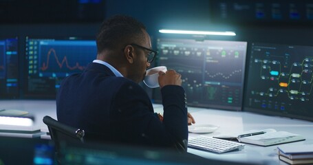 African American IT technical specialist works on computer with data server and real-time analysis charts in modern monitoring office. Team of software engineers and big digital screens on background.