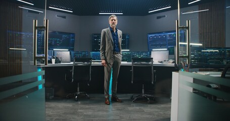 Mature big data scientist in business suit puts on glasses, stands in monitoring room and looks at camera. Multiple big screens on the wall with displayed live analysis feed at background. Dolly shot.