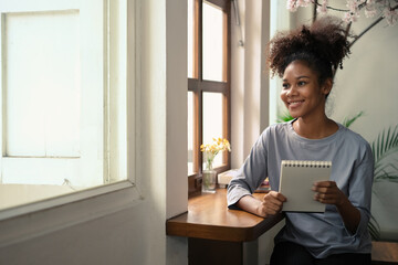 Beautiful African American woman holding notepad and looking out of window, thinking over creative ideas for research