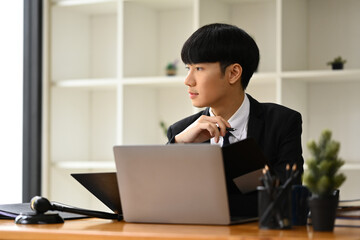 Thoughtful young businessman sitting at office desk and looking out of the window, daydreaming, visualizing future