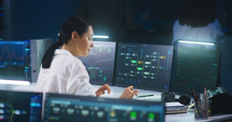 Female IT technical specialist works on computer with data server and blockchain network database in modern monitoring control room. Team of software engineers and big digital screens on background. - Powered by Adobe