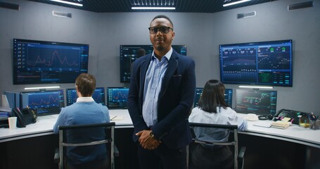 African American IT scientist stands in monitoring room and looks at camera. Multi cultural big data engineers work at computers. Multiple screens with displayed real-time analysis charts. Dolly shot.
