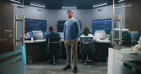 Adult IT scientist smiles, stands in monitoring room and looks at camera. Big data engineers work at computers at background. Multiple big screens with displayed real-time analysis charts. Dolly shot.