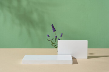 Against green background, two white podiums displayed with few lavender flowers. Stage showcase on...