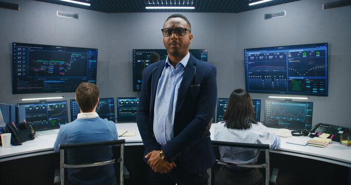 African American IT scientist stands in monitoring room and looks at camera. Multi cultural big data engineers work at computers. Multiple screens with displayed real-time analysis charts. Dolly shot.