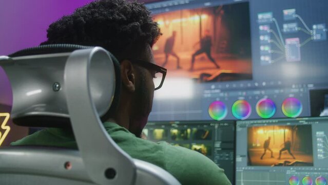 Back view of African American editor making color correction, working at home studio. Action movie footage and software interface on computer and big digital screen. Concept of film post production.