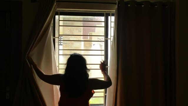 A beautiful Indian lady opening window curtains looking through window enjoying nature. Concept of hope.