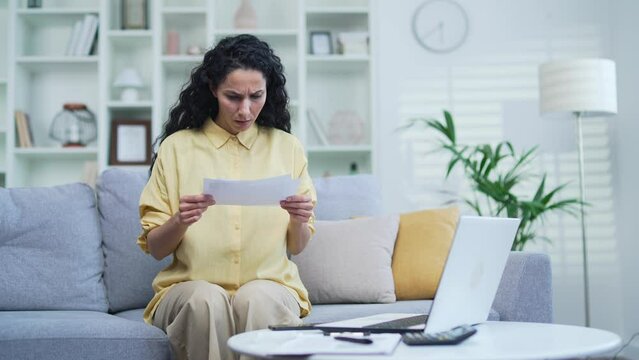 Frustrated shocked woman looking at large utility bills sitting on the sofa in the room at home. Upset female holding a paycheck, thinking about problems in the household budget and financial expenses