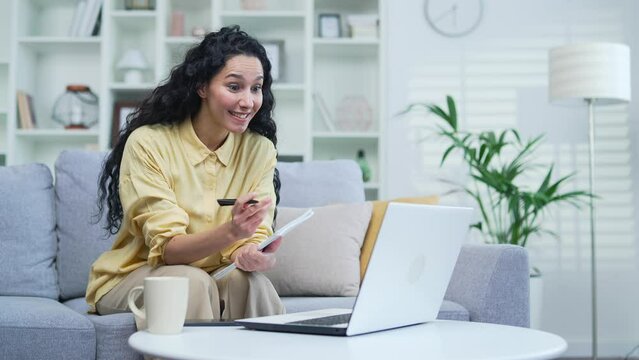 Female student listens to online e-learning of a remote teacher while sitting on the sofa at home. A woman at a distance learning seminar writes in a notebook using a laptop communicates with a tutor