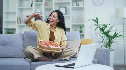 Young cheerful woman eating pizza sitting on sofa in living room. A smiling female holds a paper box in her hands and is happy about the fast delivery of food. The student ordered food to take home