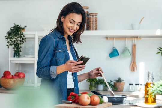 Smiling beautiful woman using her mobile phone while cooking vegetables in the kitchen at home.