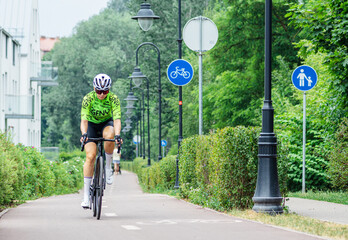 Girl with helmet cycling on the bike road with blue road sign or signal of bicycle lane among green trees and hedges, spring summer nature and street lamps