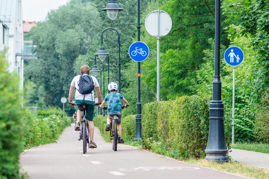 Father with backpack and son with helmet cycling together with bikes on the bike road with blue road sign or signal of bicycle lane among green trees and hedges, spring summer nature